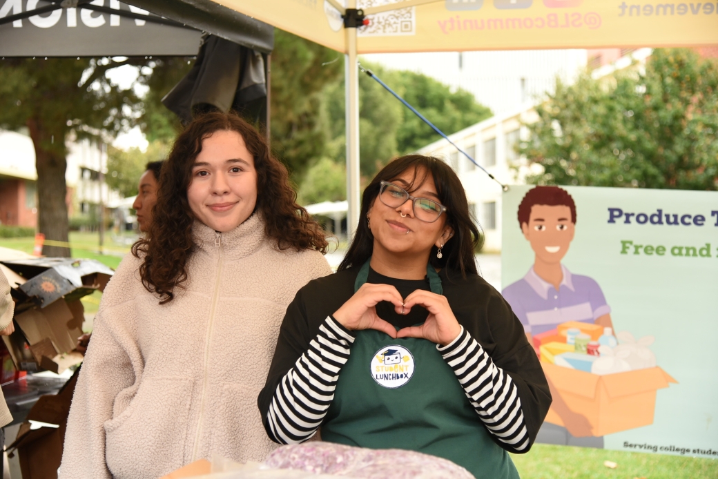 two students standing beside each other. one has short hair and a fuzzy white sweater on and another has a black long-sleeve top with a green apron on it and she is making a heart with her hands.