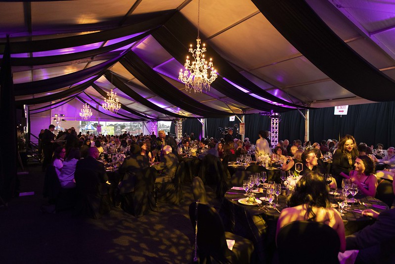 Interior shot of the alumni awards gala. Attendees sitting at round tables with drapery above, clothed in a purple light with chandeliers. 
