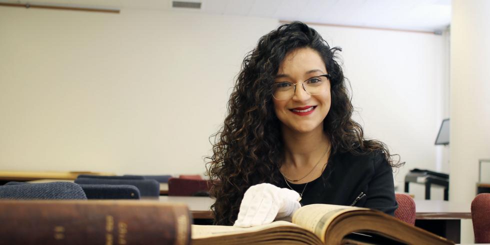Amalia Castaneda wears white gloves, with her hand in an old book, and smiles at the camera