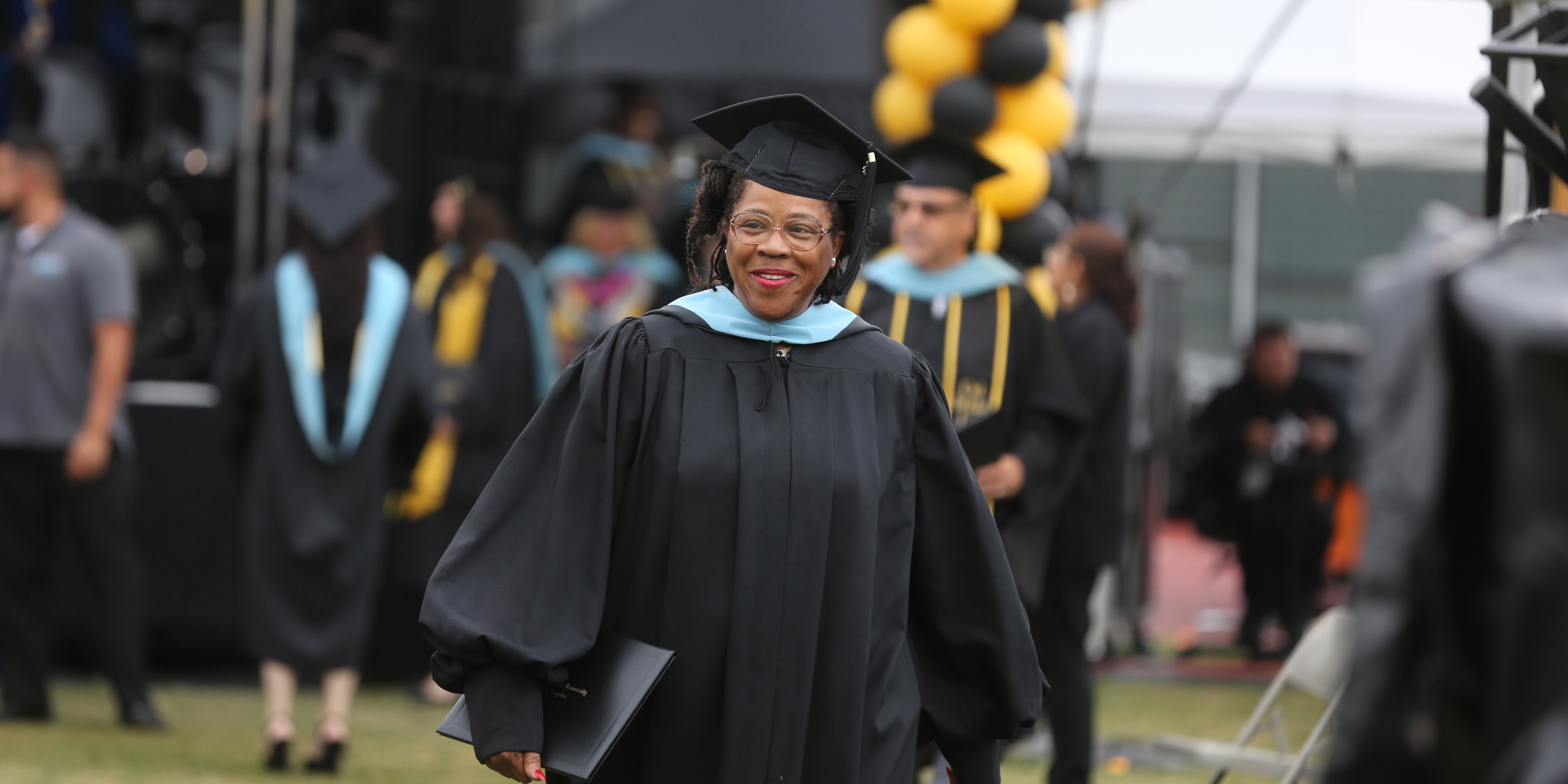 A doctoral student wears a tam, gown, and hood and carries a diploma at commemencement