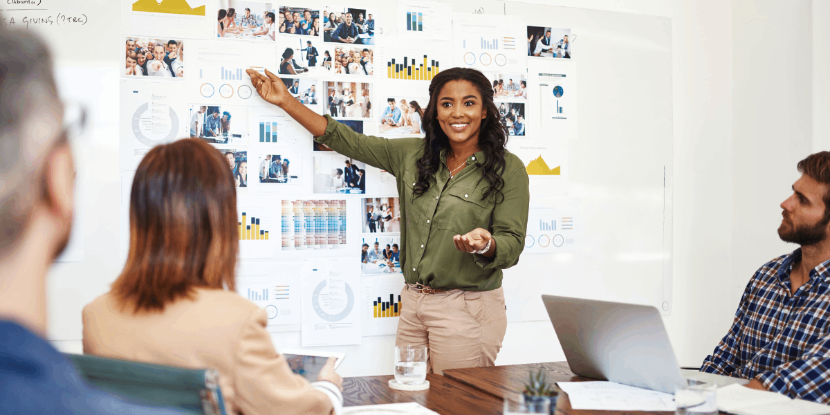 Person stands pointing at whiteboard containing post it notes and photos. Co-workers at conference table look at her.