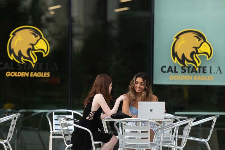Two students sitting at an outdoor patio table. Window decals on the building behind them say Golden Eagles with the mascot head