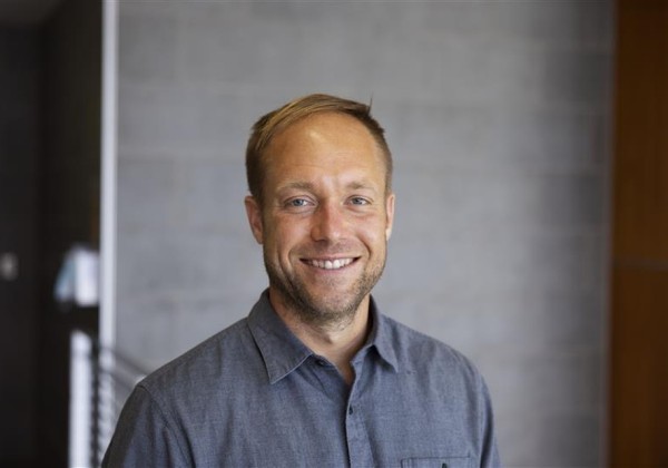 Portrait of a smiling man in a button-down shirt standing indoors against a neutral gray wall background.