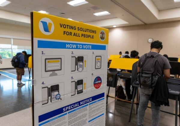 A 'How to Vote' instructional sign is posted in the foreground of a Cal State LA polling location, with students casting ballots on bright yellow voting machines in the background.