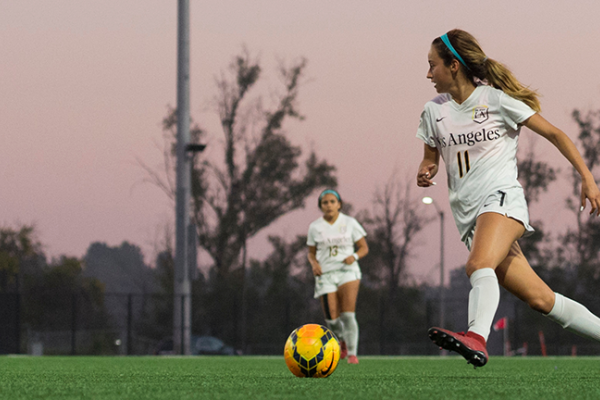Image of a member of the  women's soccer team on the pitch at dusk, pink sky behind her. 