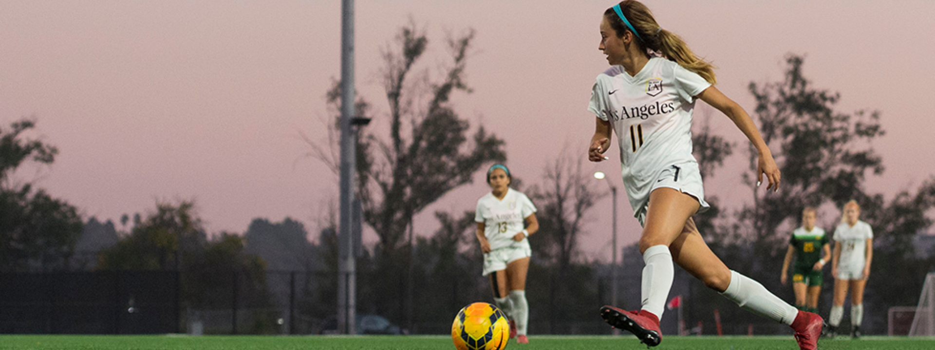 Image of a member of the  women's soccer team on the pitch at dusk, pink sky behind her. 