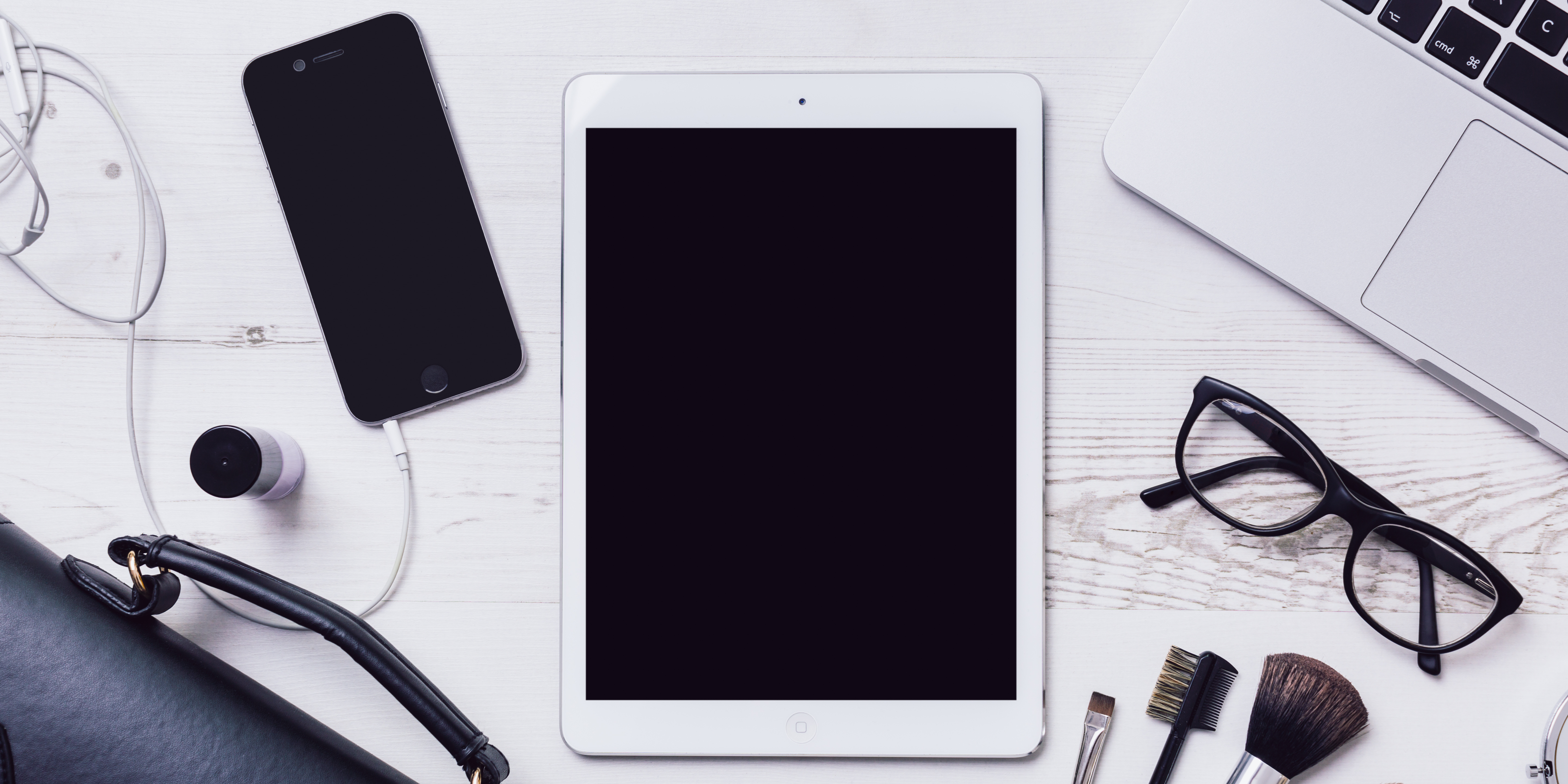 View from above of iPad, iPhone with earbuds, MacBook, glasses, and several charger cords on a desk