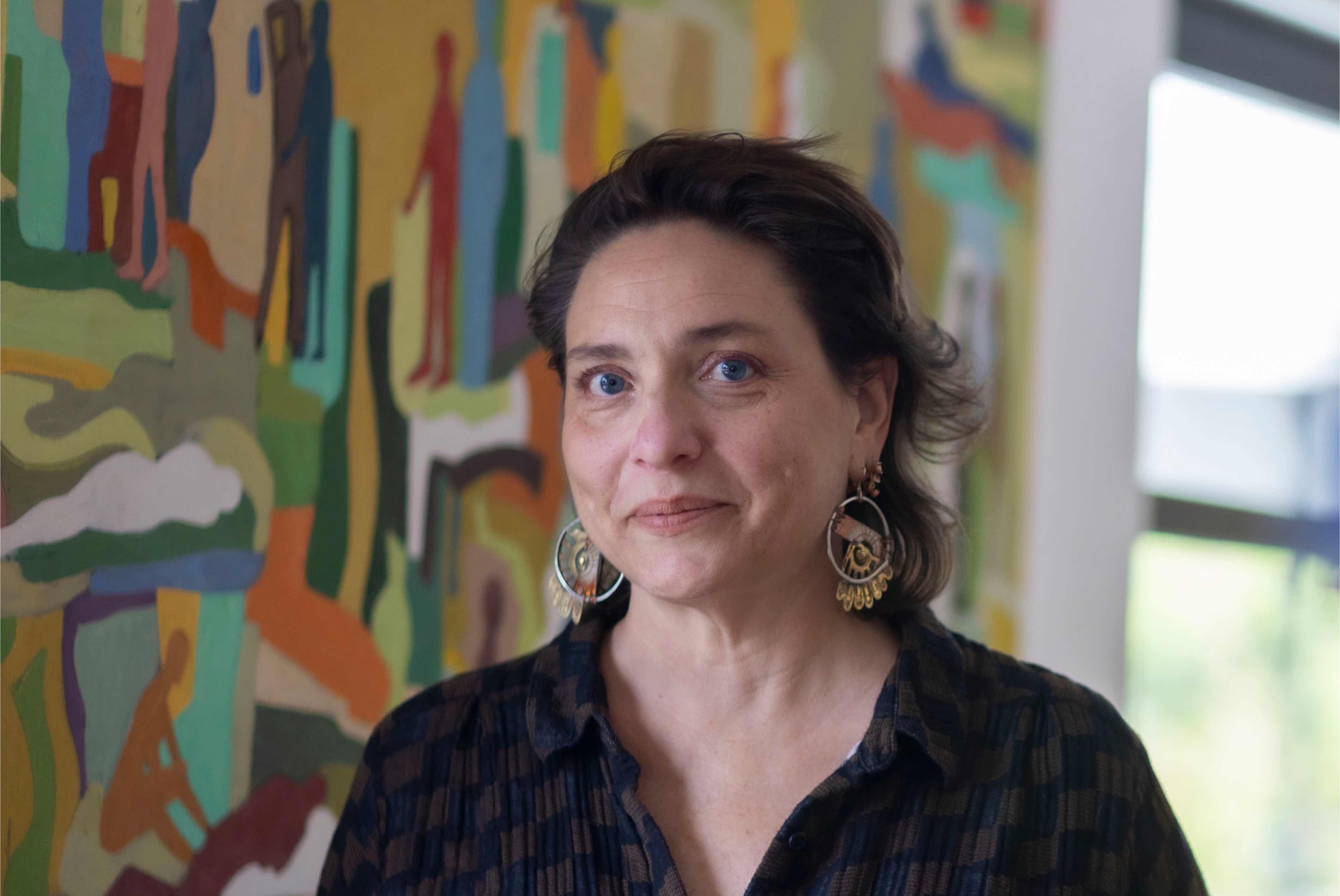 ID: Dr. Alejandra Marchevsky, short brown hair (above shoulders), blue eyes, white skin, dark blue and greyish brown checkered collared blouse with mixed gold and silver earrings and a colorful painting mural in the background