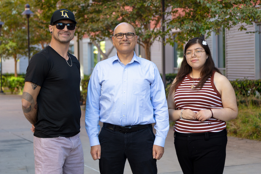 A man in a black shirt and baseball cap stands on the left, Professor Jamil Momand stands in the center wearing a light blue button-down shirt, and a student stands on the right wearing a red-and-white striped sleeveless top.
