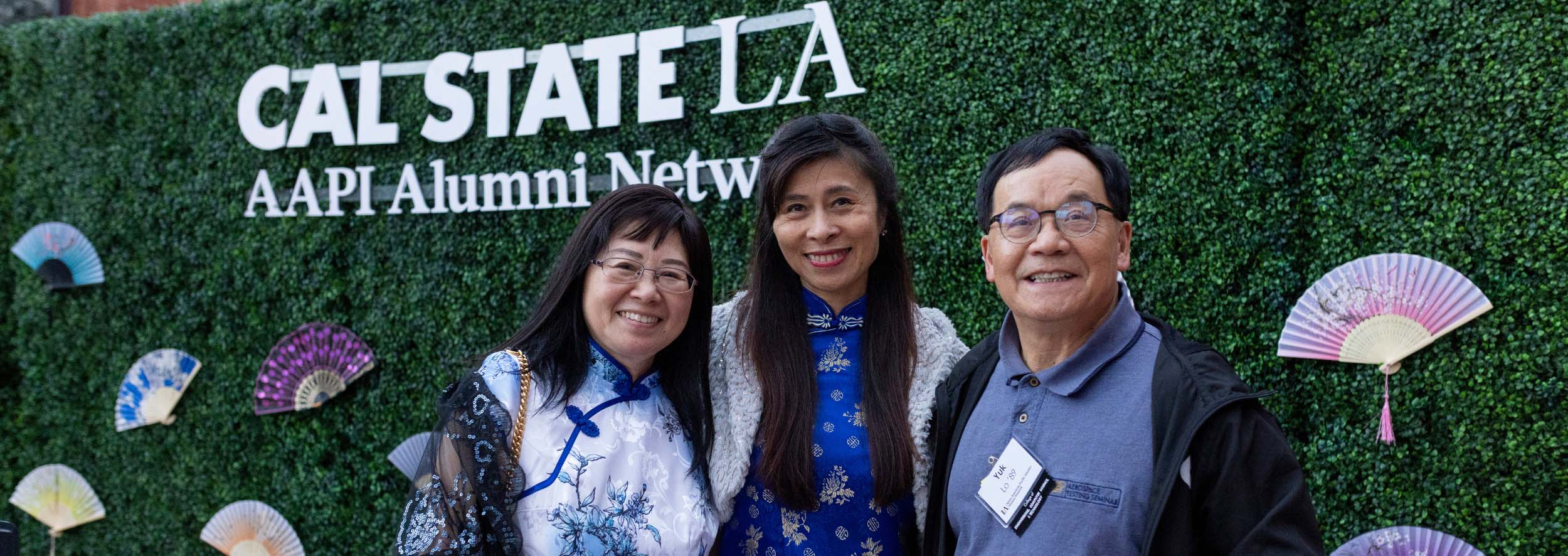 Two women and a man smiling in front of hedges with a banner that says Cal State LA AAPI Alumni Network