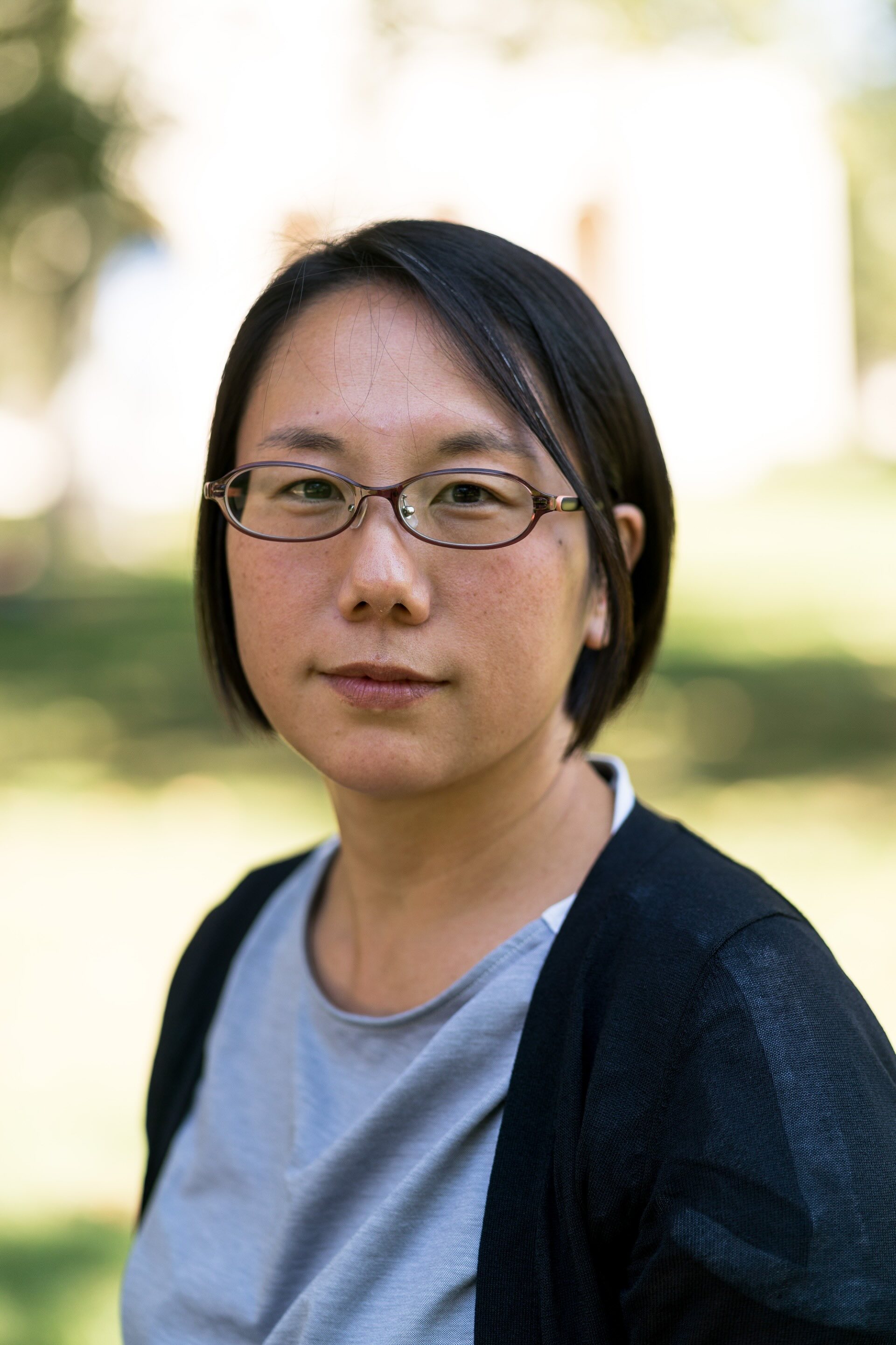 Aya Nakagoshi with short, straight dark hair and oval glasses, wearing a gray shirt and dark jacket, with a calm, neutral expression outdoors against a blurred background.