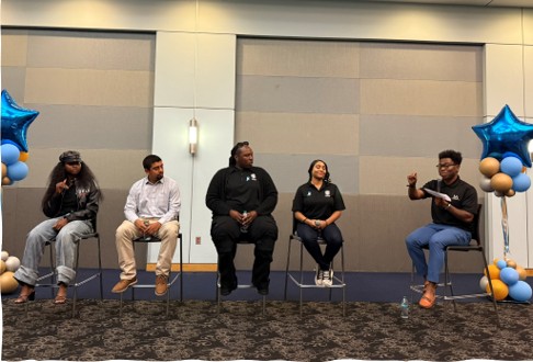 A group of four people on tall stools listening as one is speaking in the microphone