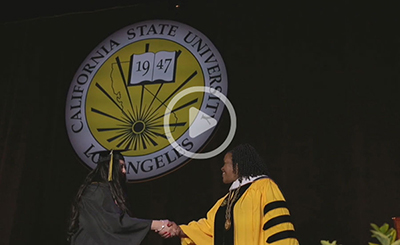 A graduate in a black cap and gown shakes hands with President Eanes on stage, with the Cal State LA seal behind them.
