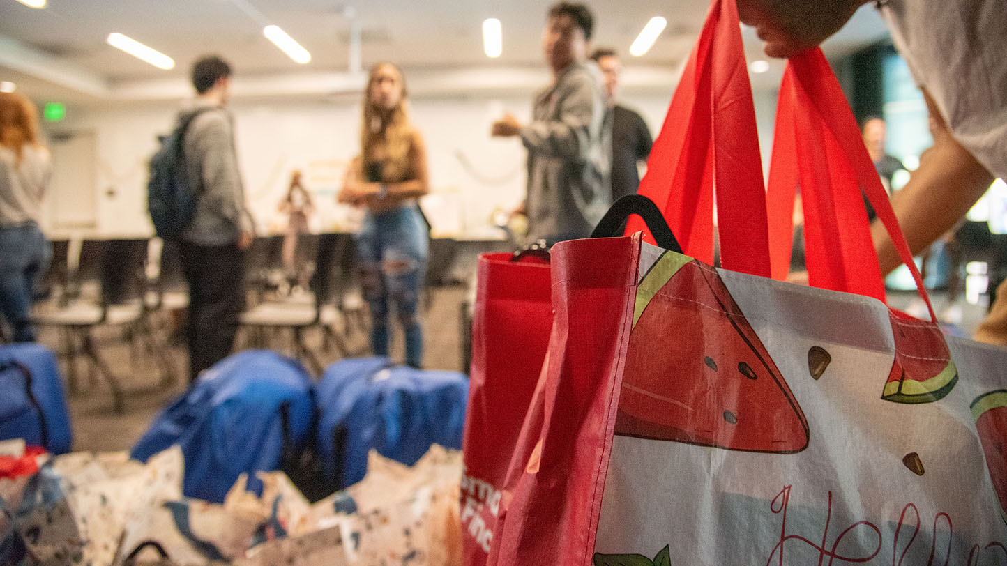 A large tote bag filled with goods. People standing near rows of duffle bags on the floor.