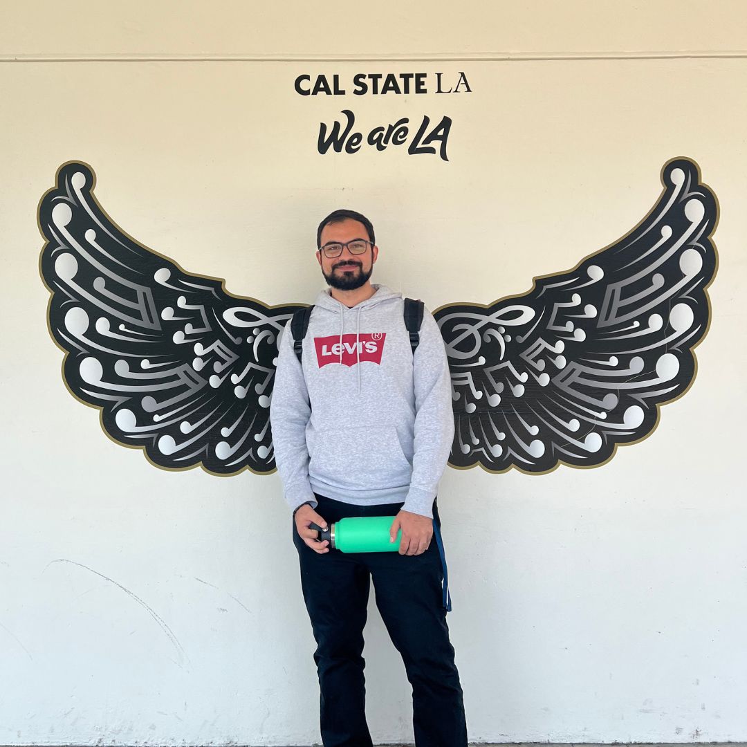 A person in a grey sweatshirt posing for a picture against black wings painted on a white wall.