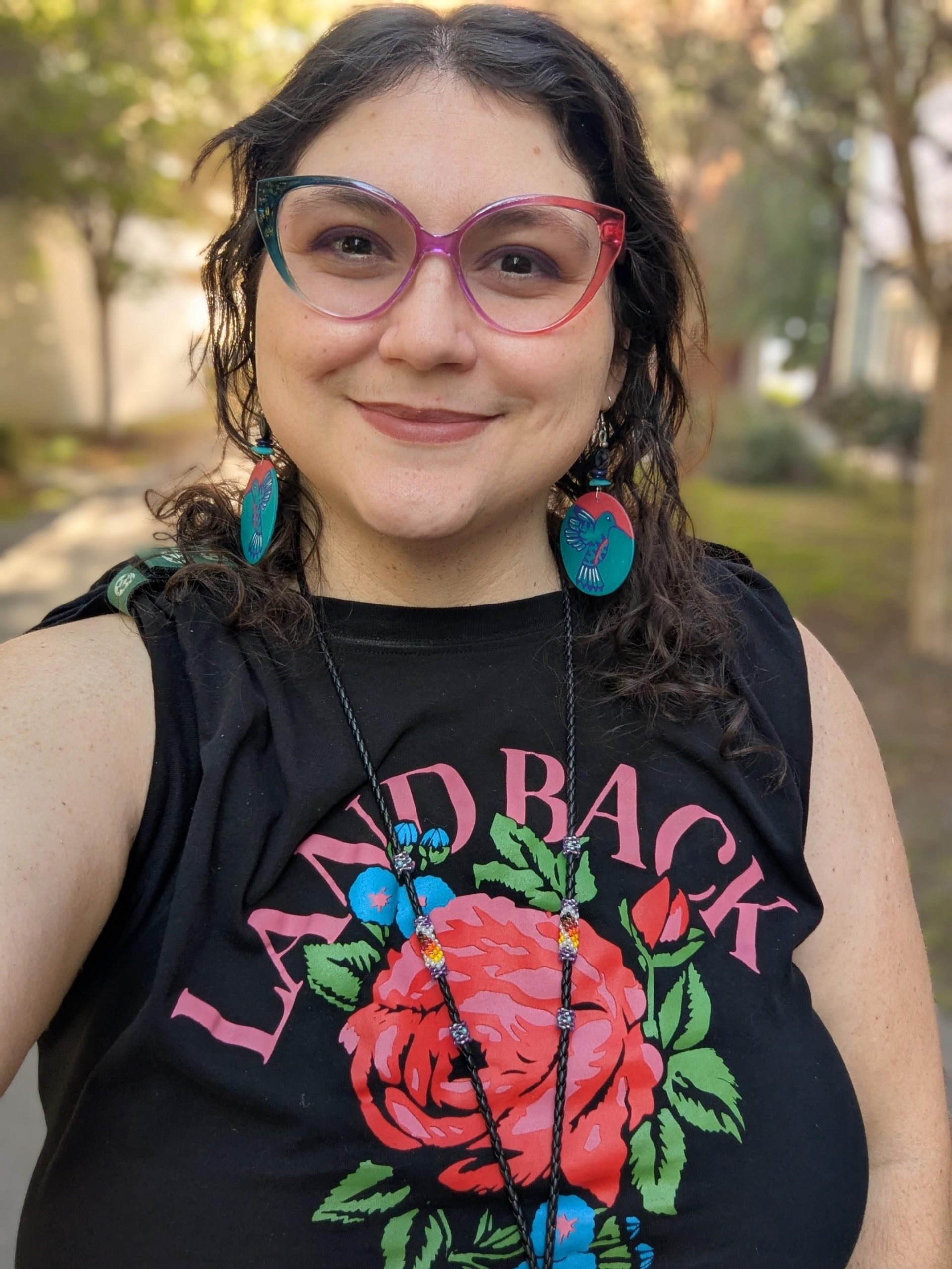 Dr. Jessica Margarita Gutierrez Masini wearing black tank top with "Land Back" text, blue earrings, dark wavy hair, rainbow glasses.