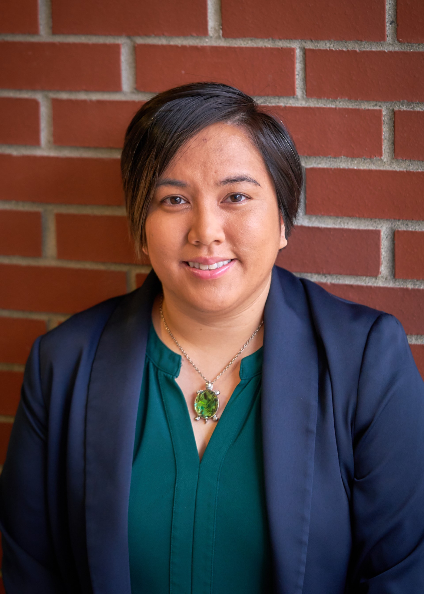 ID: Dr. Pau Abustan, brown eyes, beige skin, short black hair, smiling, wearing a navy blazer, green shirt, green abalone turtle necklace with red brick background