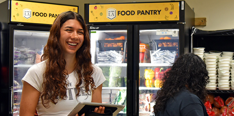 A person with long hair smiling, holding a clipboard while standing in a room with two large refrigerators that have Cal State LA Food Pantry panels at the top.