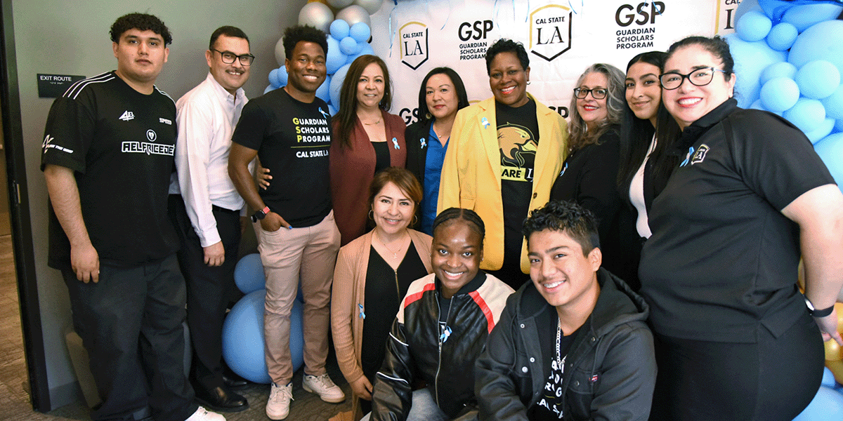 A large group of staff and students, standing with the university president, in front of a step and repeat framed by balloons.