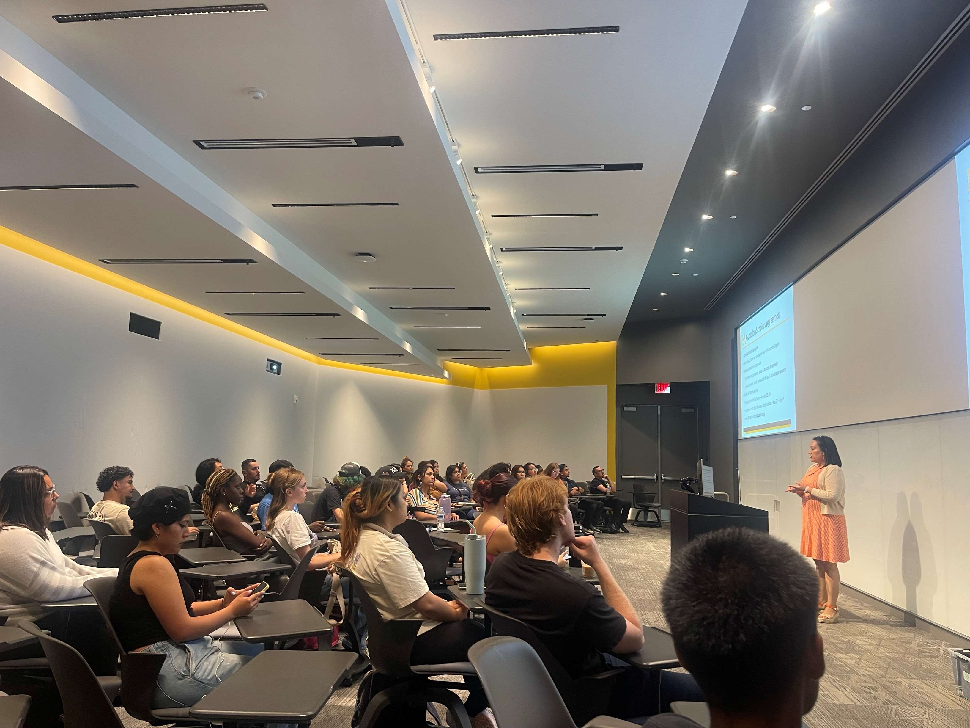 A room full of students with a woman in a peach color dressed speaking to them.