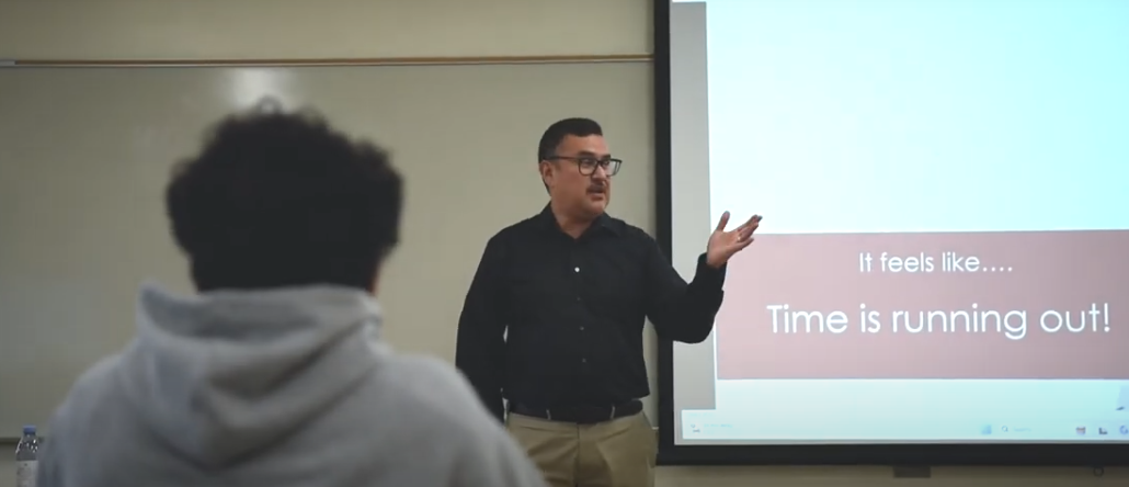 Photo of man in black shirt, glasses, and tan pants giving a presentation to a student in a grey hoodie.