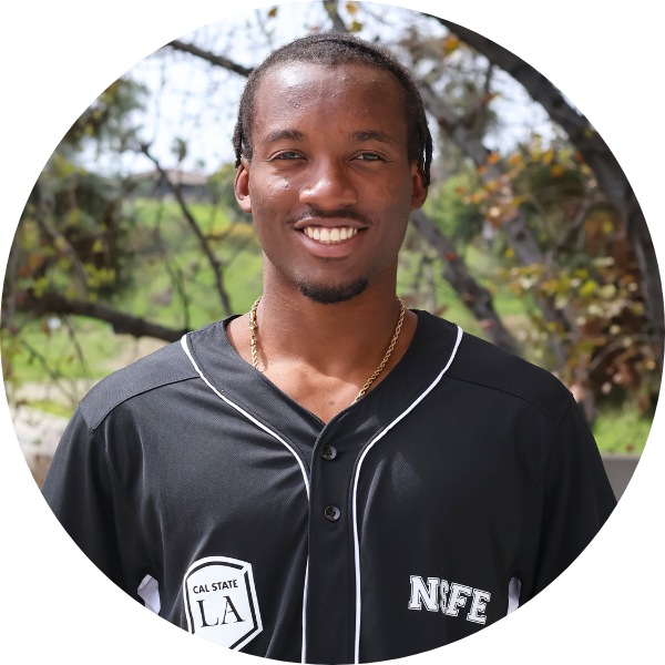 A man with a black baseball jersey smiling toward the camera. Trees in background.