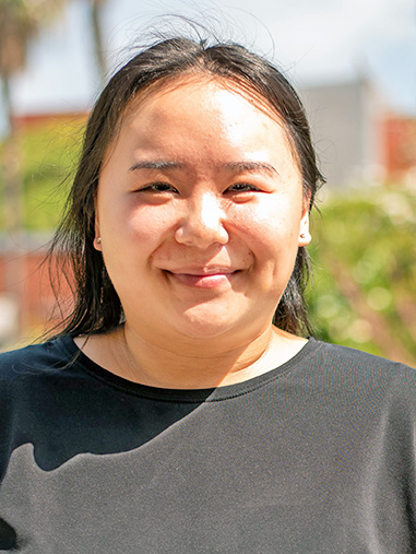 A person with long hair styled back, outdoors in the sun.