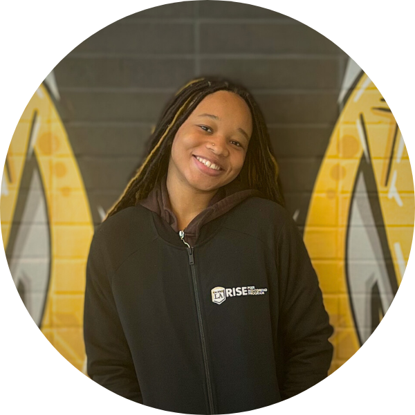 A woman with a black jacket smiling in front of a black wall with yellow markings.