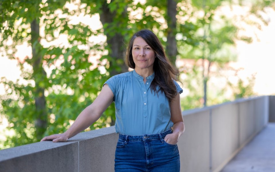 Student wearing a blue top and jeans standing outside with trees in the background
