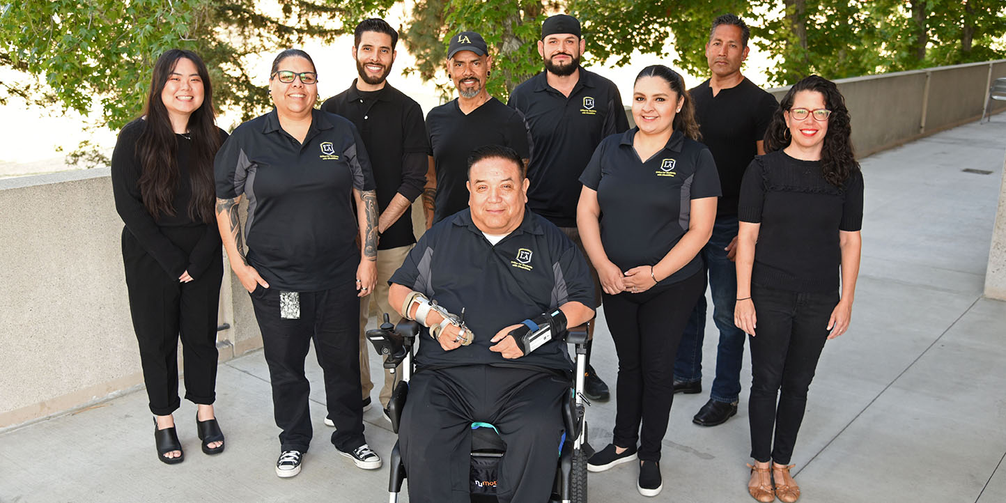 Nine people wearing matching shirts standing in a shady areas outdoors, smiling.