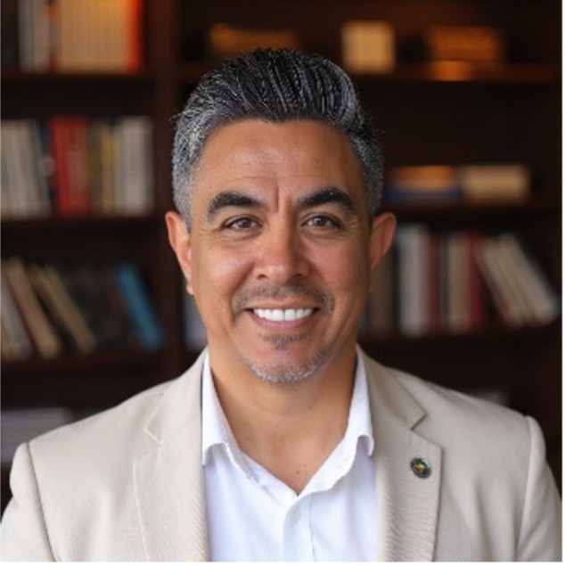 Ozzie Lopez wearing a tan blazer, a white dress shirt.  He's standing in front of a book of shelves
