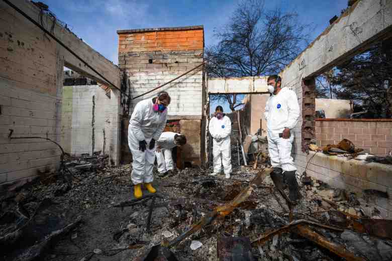 A group of archaeologists and volunteers, dressed in white protective suits and masks, carefully search through the charred remains of a burned-down home.