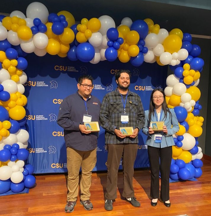Three students (2 male, 1 female) standing with their awards on a stage that is decorated with balloons