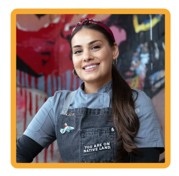 Smiling woman in a gray chef’s uniform and apron stands in front of a colorful graffiti wall, her hair pulled back with a red headband and a small pin visible on her apron.