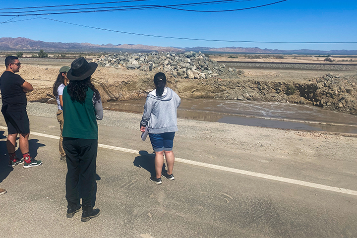 A small group of students stands near a dirt embankment observing a muddy water flow site surrounded by desert terrain and distant mountains under a clear blue sky.