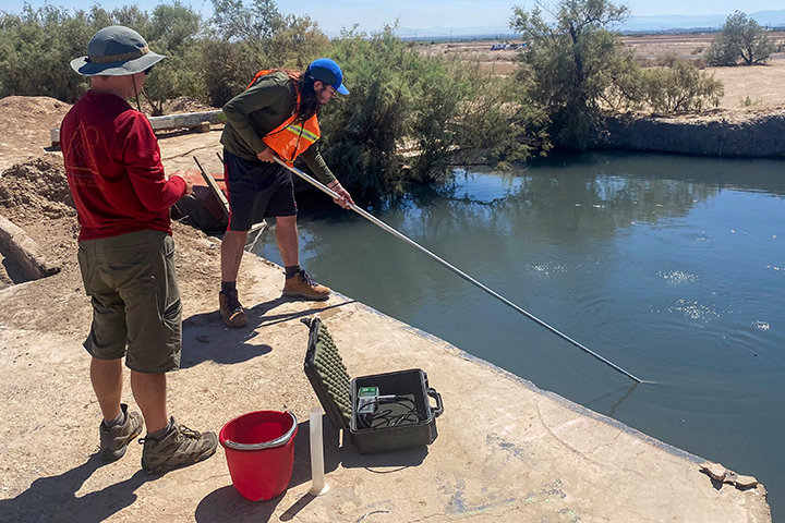 Two researchers collect water samples from a canal using a long pole. They stand on a concrete embankment beside monitoring equipment and a red bucket under bright desert sunlight.