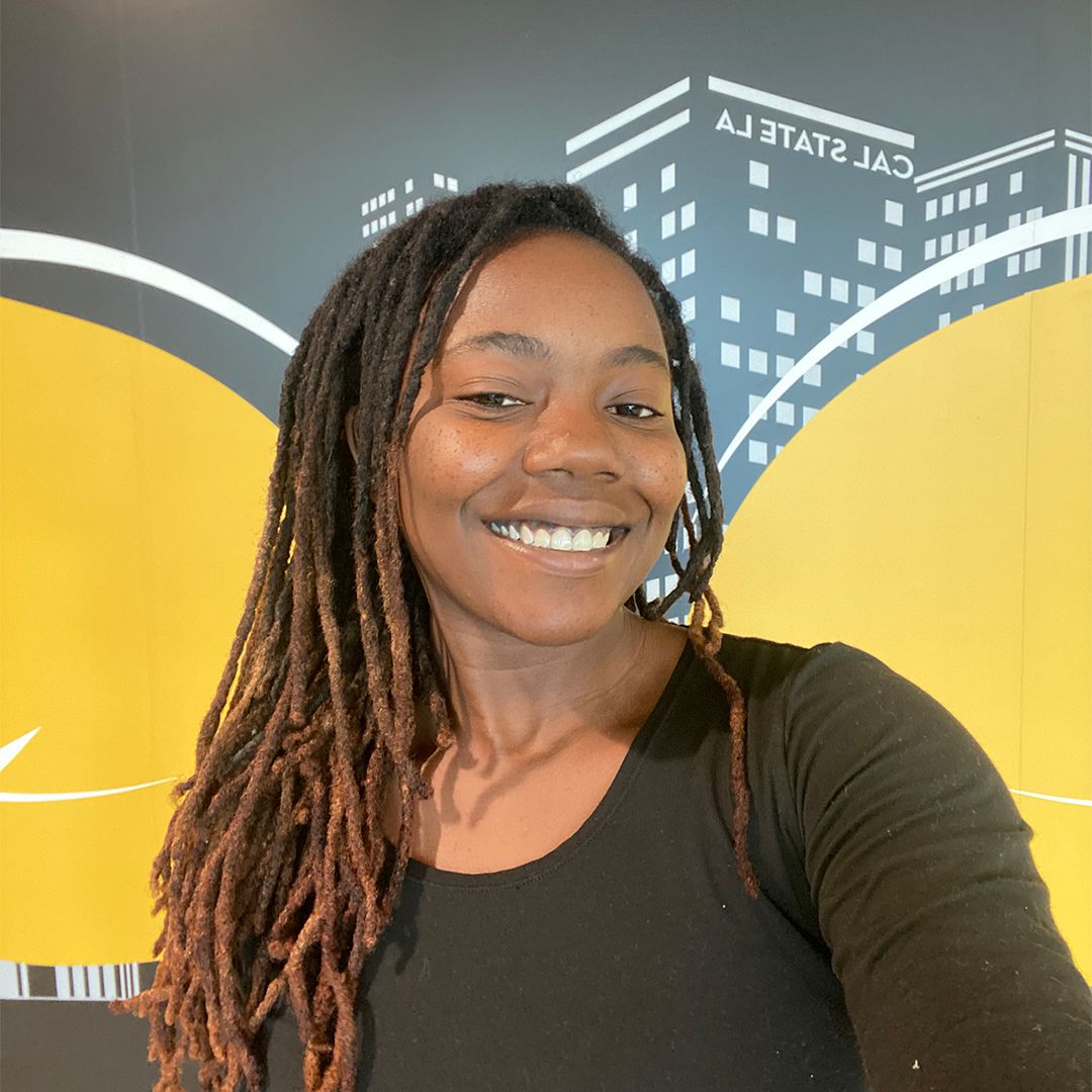 An individual with brown hair smiling against a yellow and black wall.
