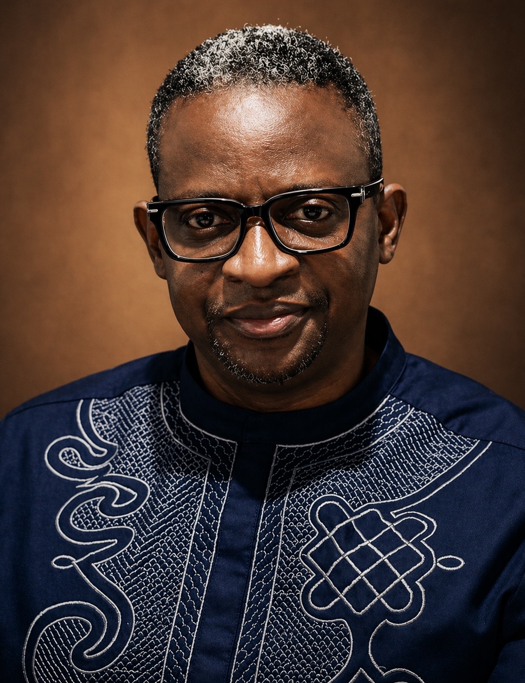 Professional head-and-shoulders portrait of a person wearing black rectangular glasses and a navy blue embroidered traditional-style shirt with intricate white patterns. They are looking directly at the camera with a slight smile. The portrait is set against a warm brown gradient background with soft studio lighting.