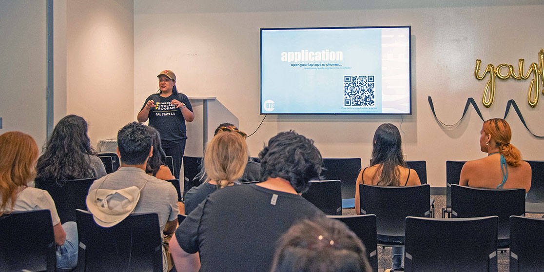 Rearview of people seated facing a person leading a presentation at the front of a room.