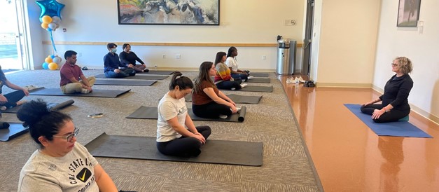 People sitting on floor in yoga position facing woman in black attire with long hair and glasses
