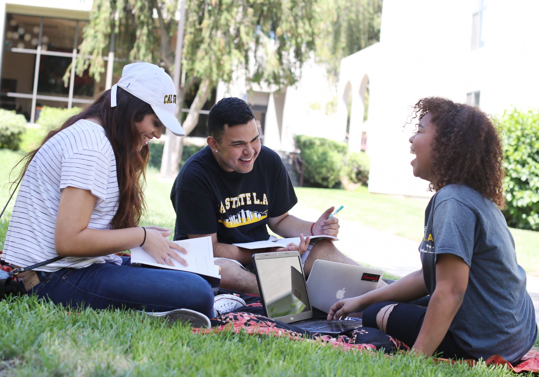 Three students sitting outdoors on a lawn, studying and smiling.