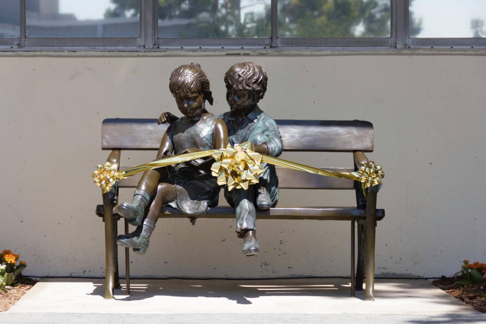 A bench with metal statues of children sitting. The bench is wrapped in gold ribbon and bows. 