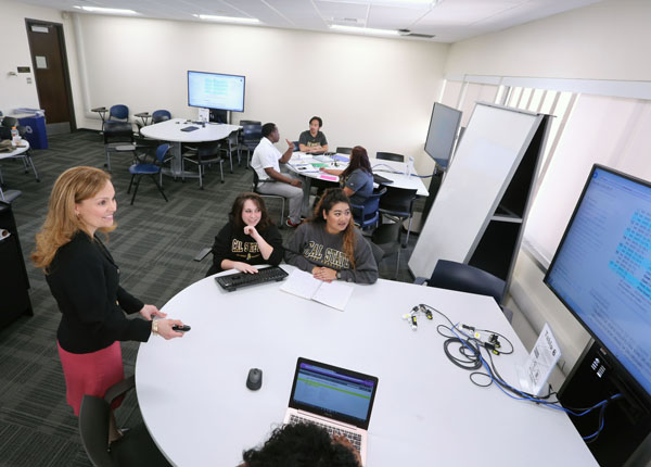 A modern classroom with computer stations and a professor giving a lecture through the available technology.
