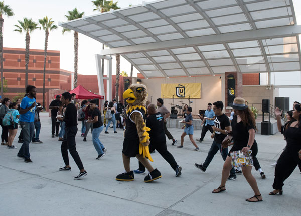 Students accompany the university mascot in front of a plaza stage.