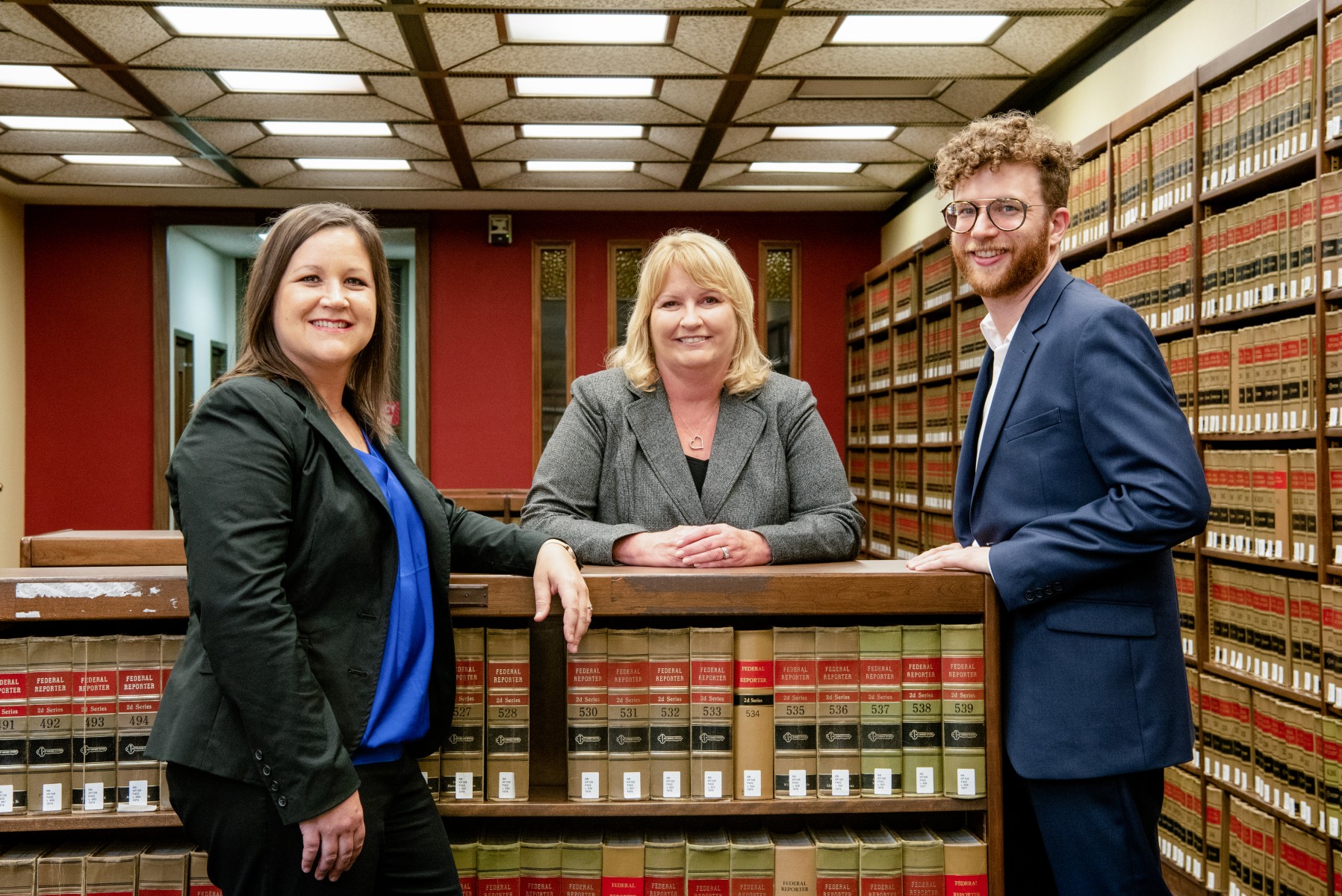 2 women and 1 man are in a law library surrounding by stacks of books. They are posed in front of and in back on a shorter book case.