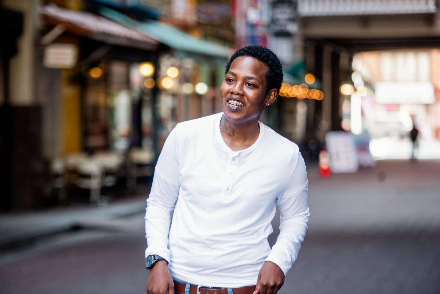 Female Black student on a street in DTLA. She is wearing a white henley, has very short brown hair, hands in pockets and smiling.