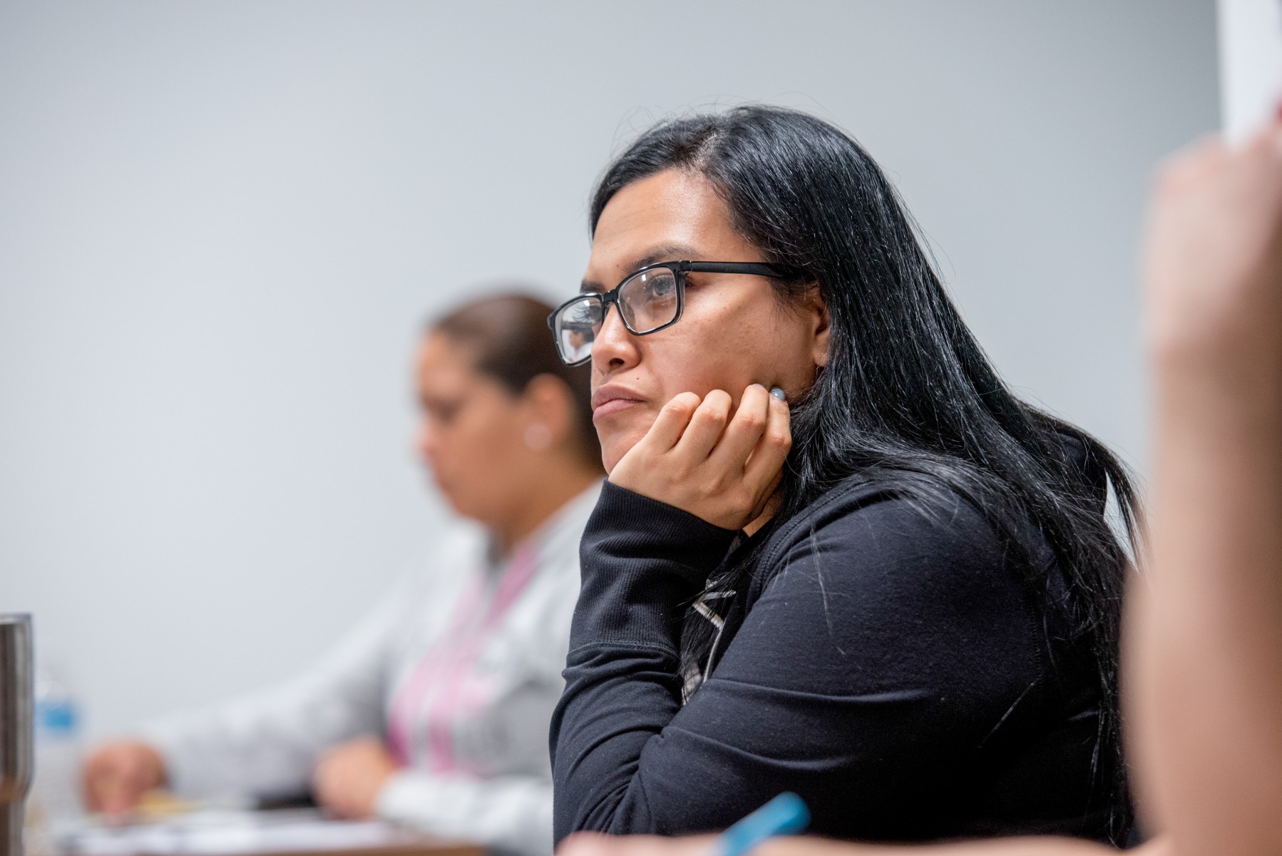 Female students with long black hair and glasses, hand on chin, looking forward, seated at desk, looking forward at instructor.