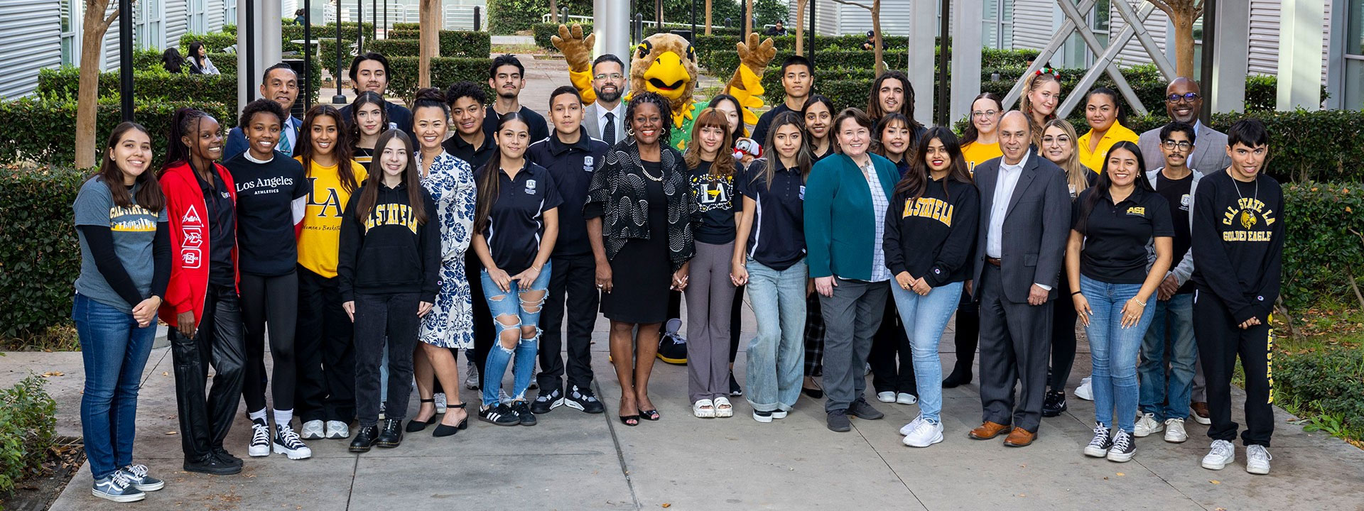 Group photo of Cal State LA students, faculty, staff, and campus leaders posing outdoors with the university’s Golden Eagle mascot. The group wears a mix of Cal State LA apparel and professional attire, standing together on a campus walkway surrounded by trees and landscaping.
