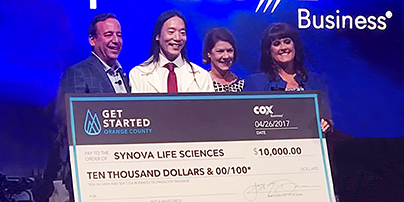 Alum John Chi holds giant check from Synova Life Sciences for $10,000. He is surrounded by three people.