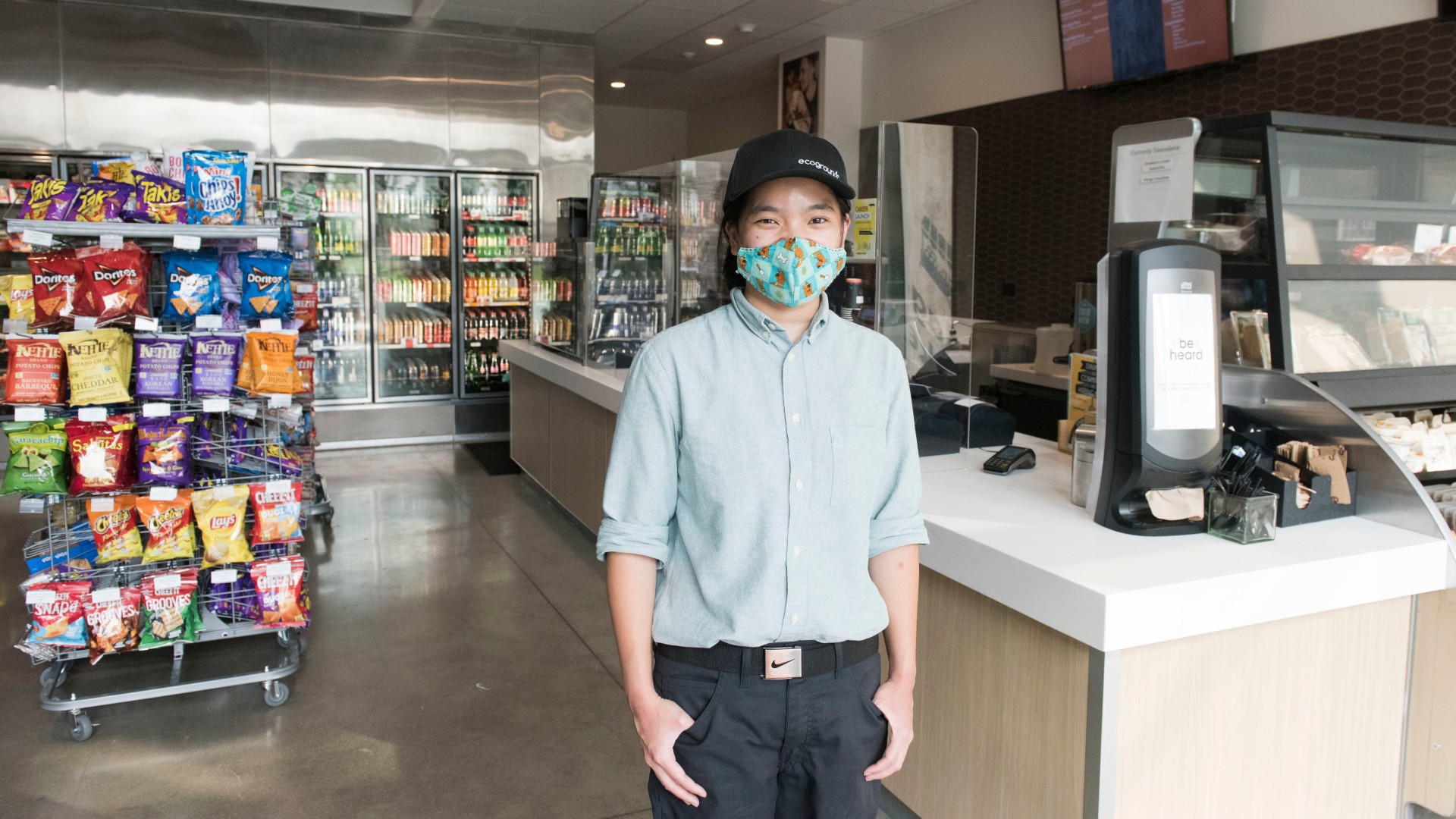 Person with a baseball cap and a facial covering poses for a photo in a convenience store.